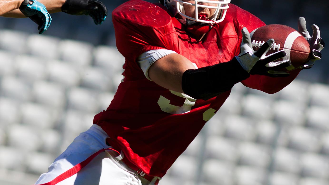 College football wearing uniform, helmet, and arm sleeve quarterback catching the ball. 
