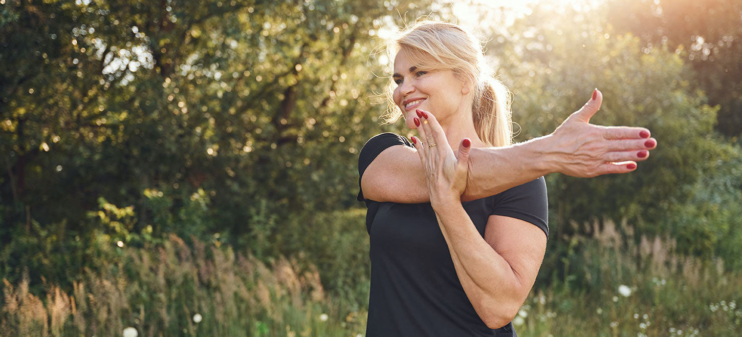 Blond middle-aged woman doing cross-body shoulder exercise in nature