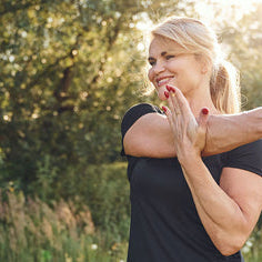 Blond middle-aged woman doing cross-body shoulder exercise in nature